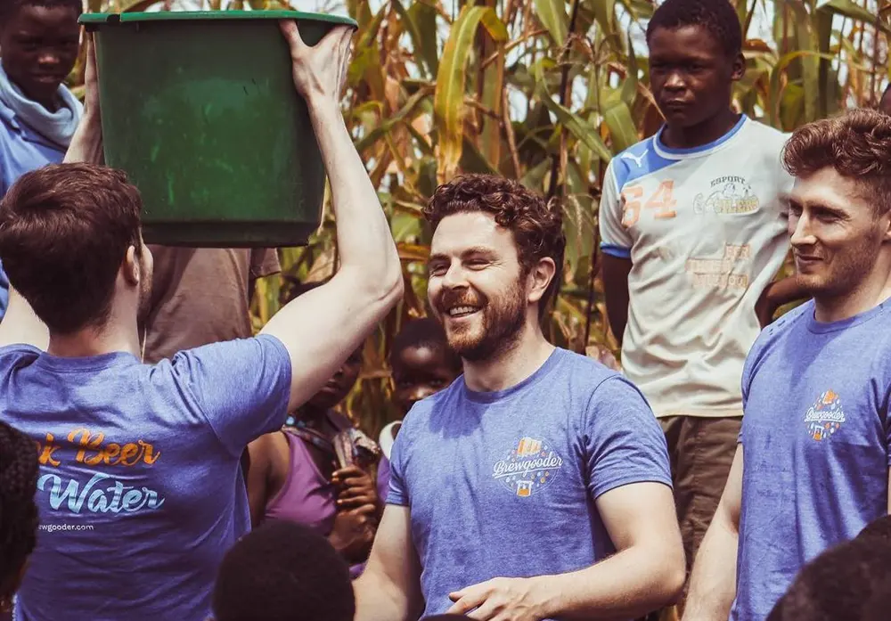 Brewgooder founder Alan Mahon smiling in a Malawi maize field as team members in Brewgooder tees pour clean water